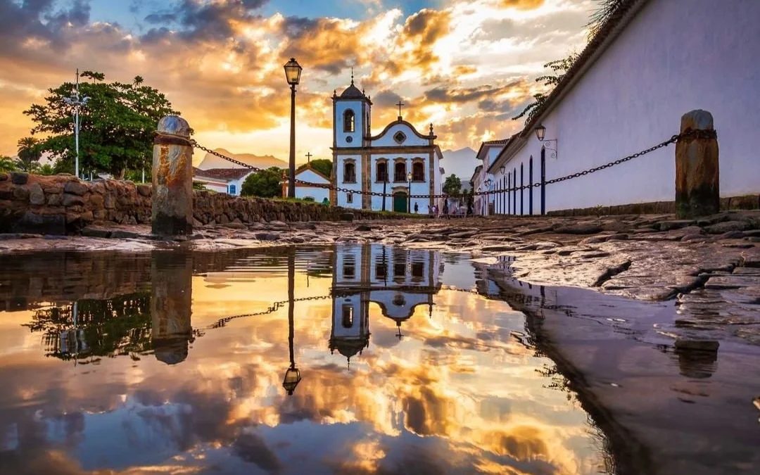 Igreja colonial no centro histórico de Paraty ao entardecer, refletida numa poça sobre o calçamento de pedra